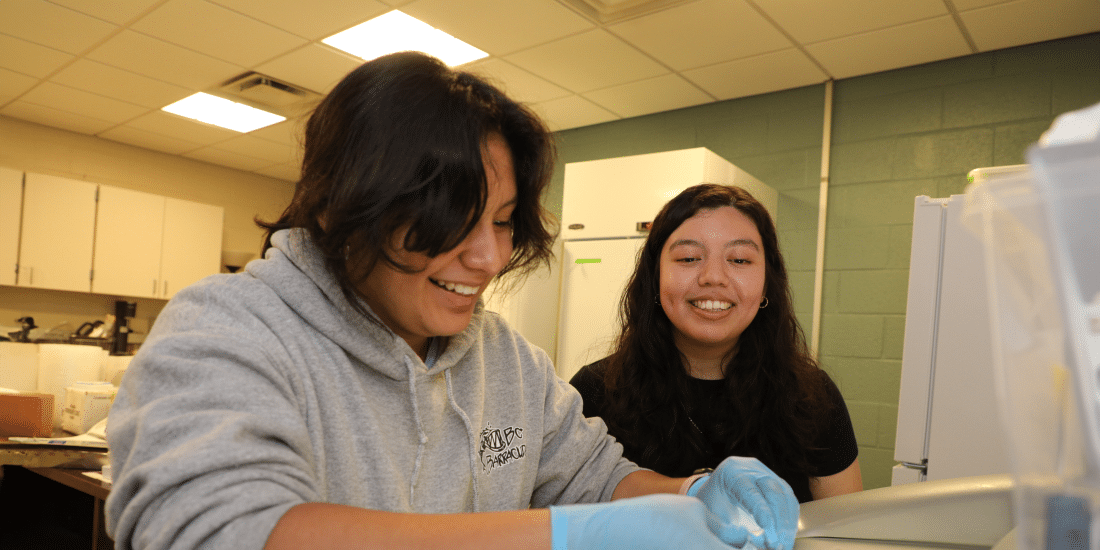 Two RMC students in a lab setting, one wearing gloves handling equipment, smiling and engaging with each other.