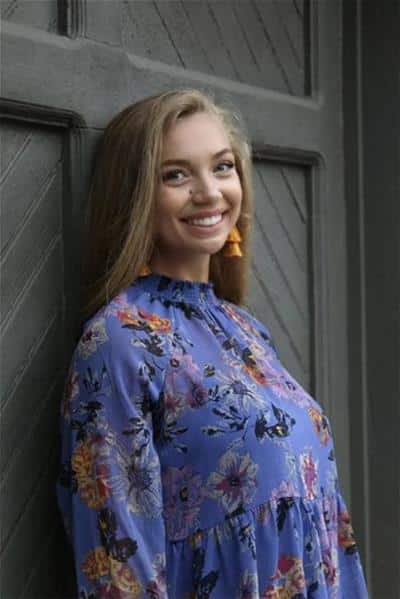 A headshot of Gracyn Draney, wearing a purple top with flowers on it. She leans against a dark gray wooden door behind her.