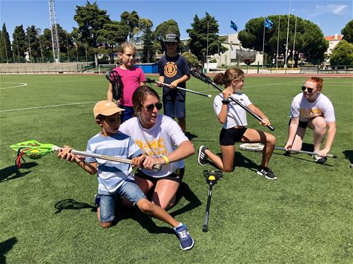 Two members of R-MC's Women's Lacrosse team, crouching alongside two youth, demonstrate how to hold the lacrosse stick.