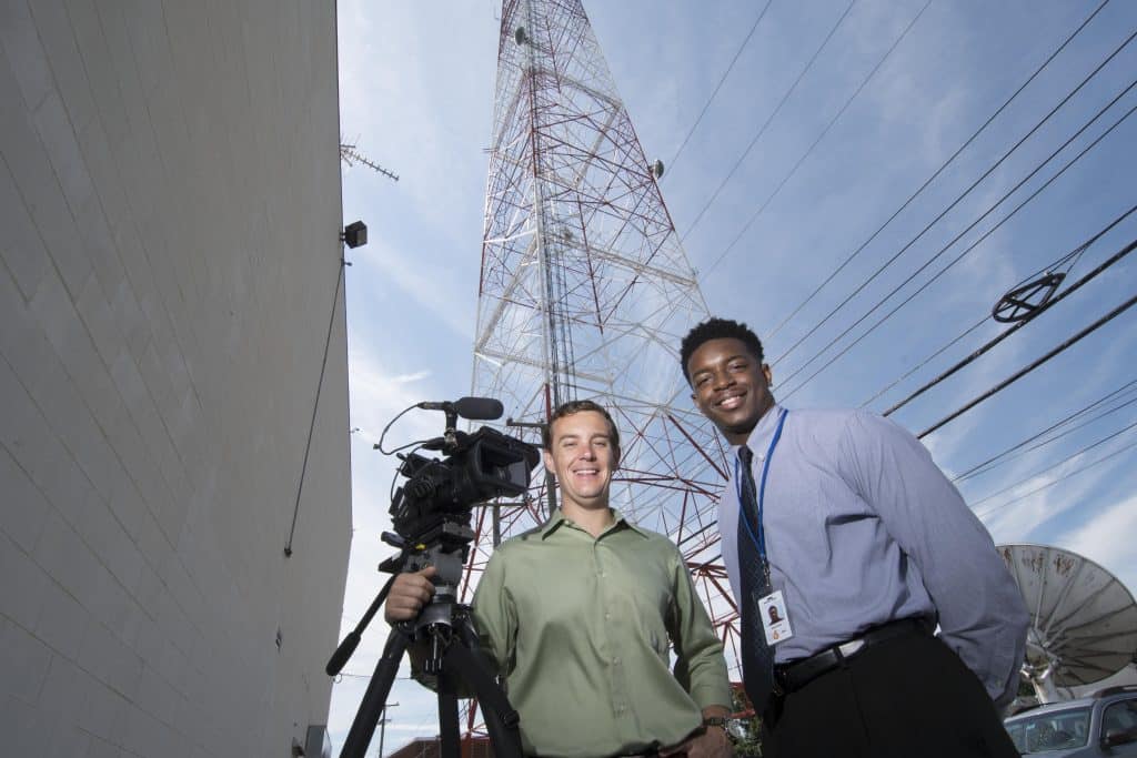 Two journalism students smiling with a video camera on a tripod