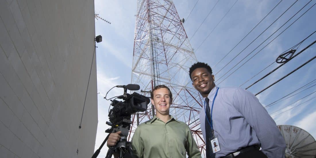 Two journalism students smiling with a video camera on a tripod