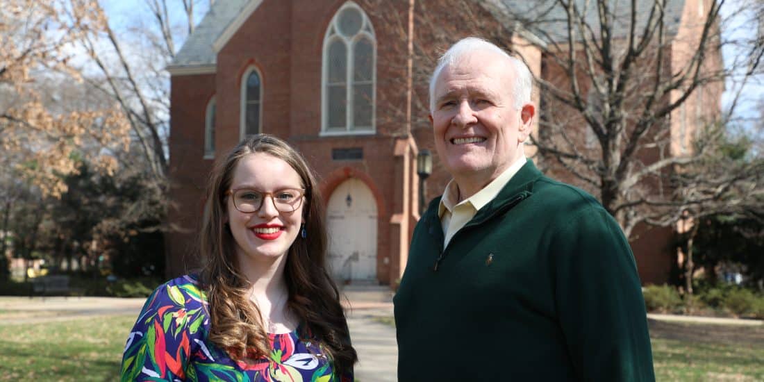 A young woman and an older man, both alumni, stand smiling in front of a brick church on a sunny day. The woman wears a colorful dress, and the man dons a green sweater.