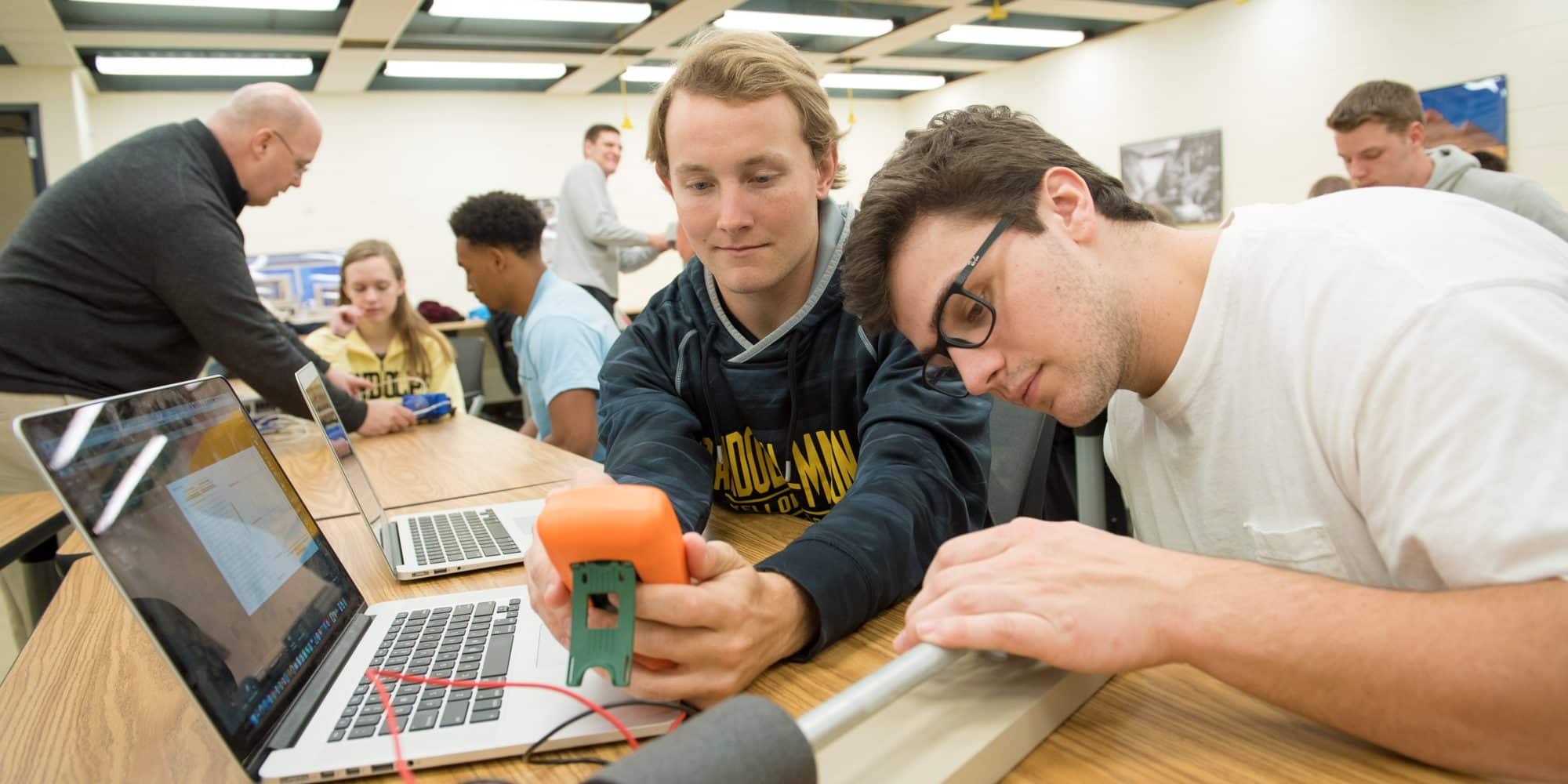 Two engineering students with a laptop and measuring equipment in a classroom while a professor supervises students in the background.