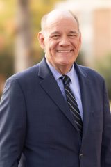Grant Azdell smiles outdoors with trees and a building blurred in the background.