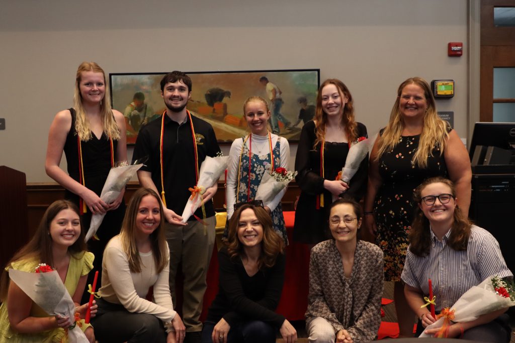 A group of nine people, some standing and others kneeling, holding flowers and smiling in a room with a painting and a door in the background.