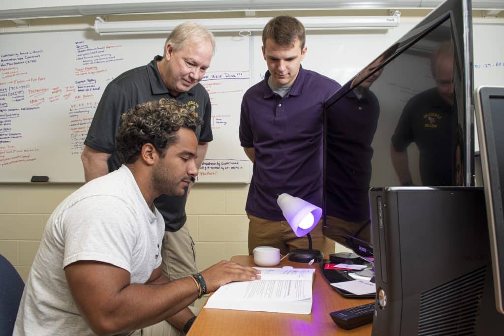 A professor and an RMC Cybersecurity student reviewing a text book at a desk in front of a computer monitor