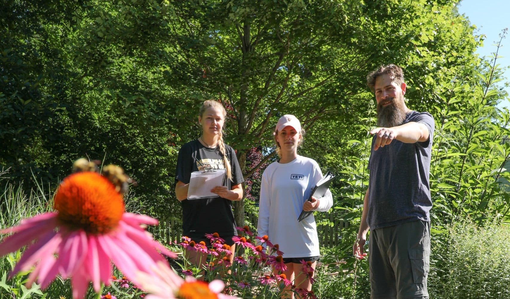 Professor Nick Ruppel pointing out at a flower with bees on it as two students take notes
