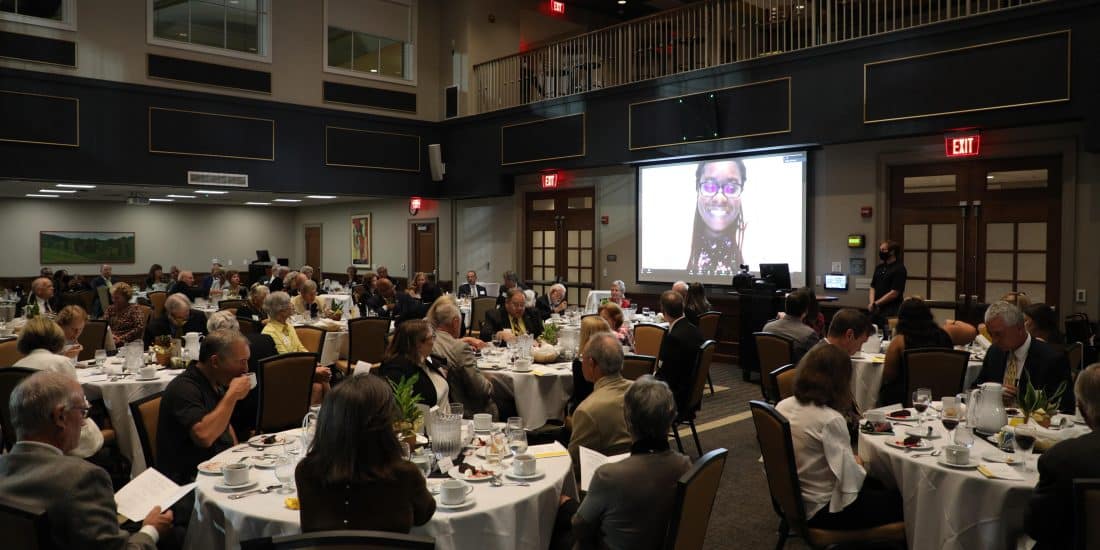 A projector screen in birdsong hall showing Jazmine "Jaz" Battle Funes surrounded by tables and chairs with people dining at the annual society of alumni dinner