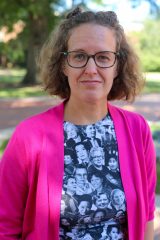 Stephanie S. Coster stands outdoors in a bright pink cardigan and photo-print shirt, with trees and a path in the background.