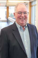 Steven D. Lang smiles while standing indoors in a well-lit hallway, dressed in business-casual attire.