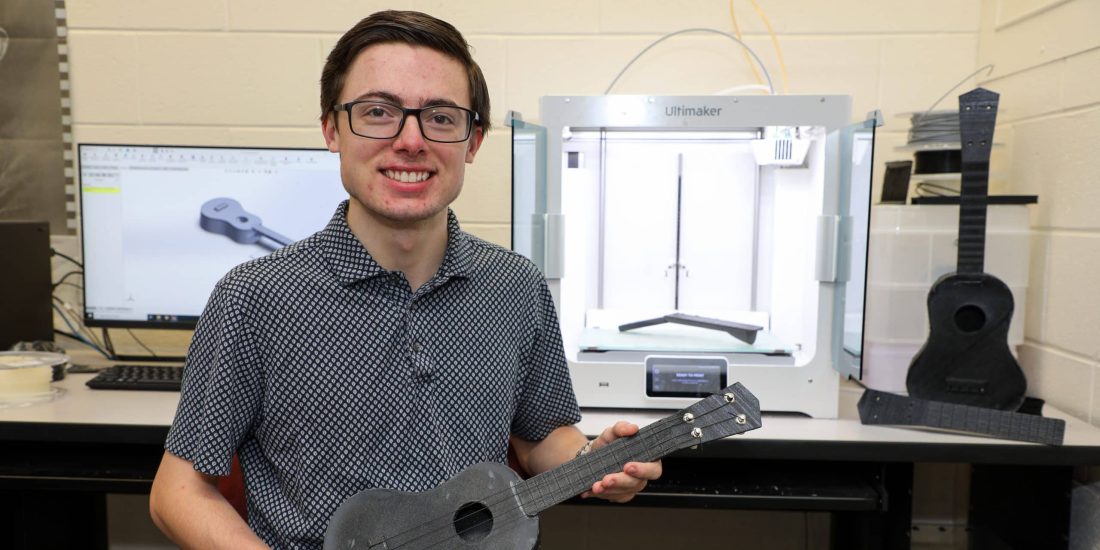 A person holding a 3D printed ukulele sits in front of a 3D printer and a computer displaying an engineering-inspired ukulele design.
