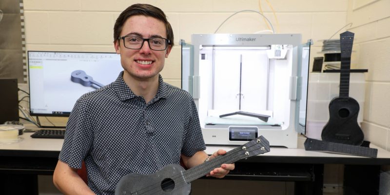 A person holding a 3D printed ukulele sits in front of a 3D printer and a computer displaying an engineering-inspired ukulele design.