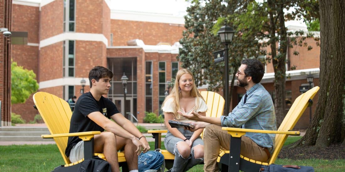Professor Volpicelli holds a book and sits two Randolph-Macon students outside on Adirondack chairs