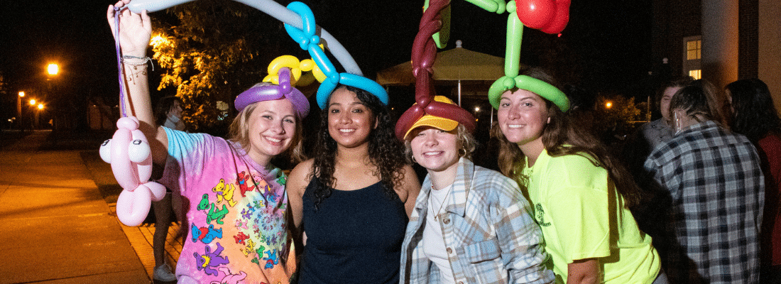 Randolph-Macon students smiling at a nighttime outdoor event, each wearing vibrant outfits and colorful balloon hats.