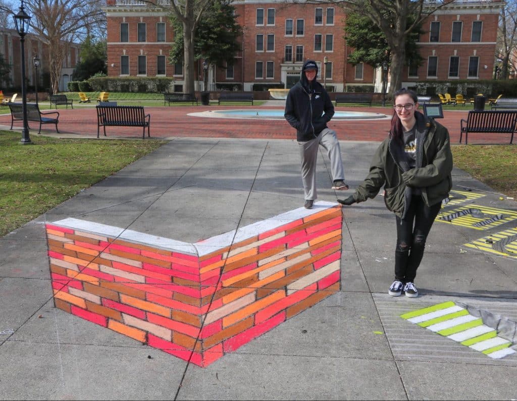 Psychology students smile near their chalk artwork designed to look three-dimensional