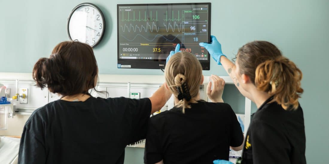 Three Randolph-Macon College nursing students face a monitor with a patient's vital signs in the RMC nursing simulation room.