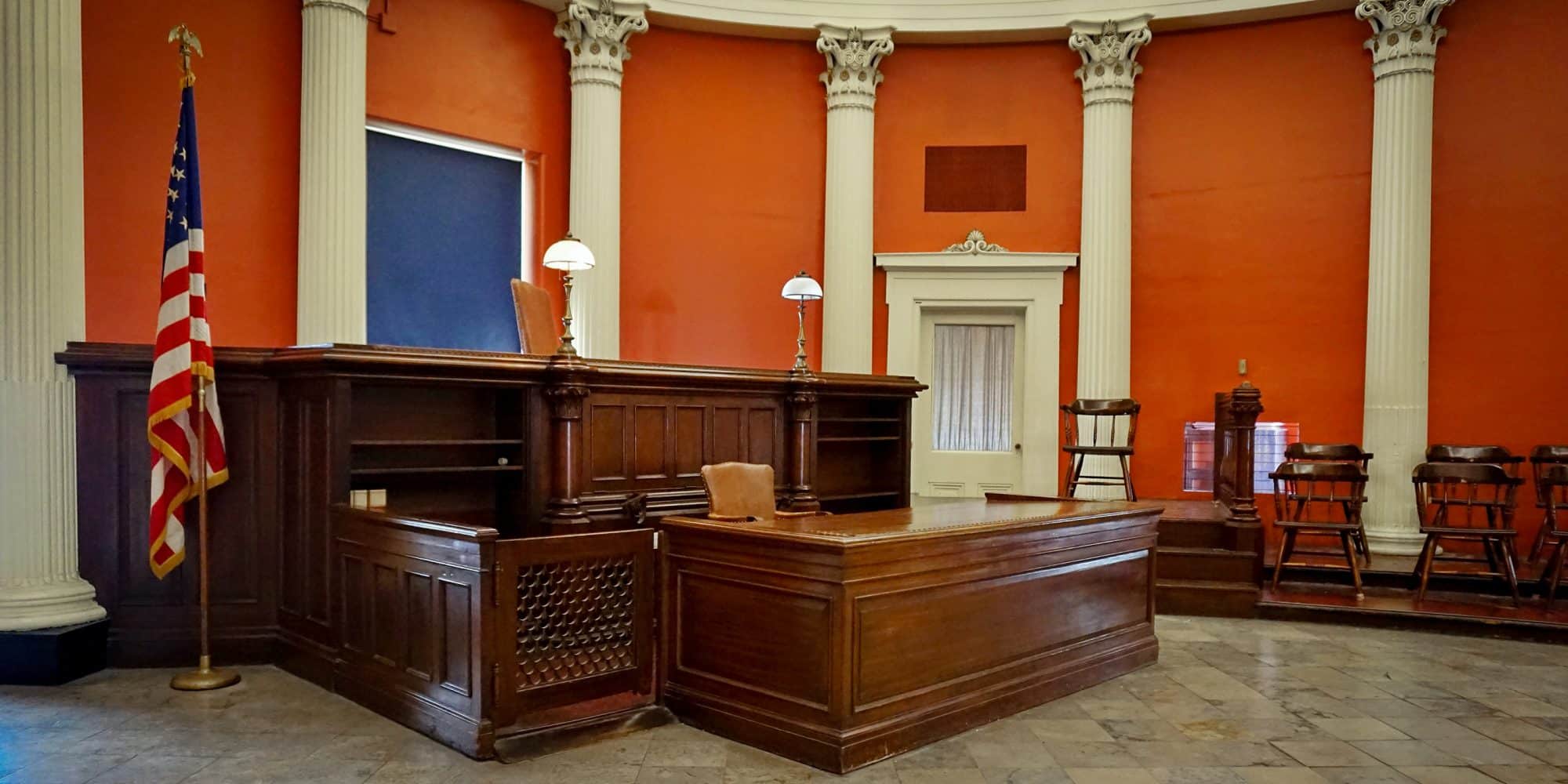 Interior of a traditional courtroom with a wooden judge's bench, an American flag, and Corinthian columns against orange walls, ideal for legal studies.