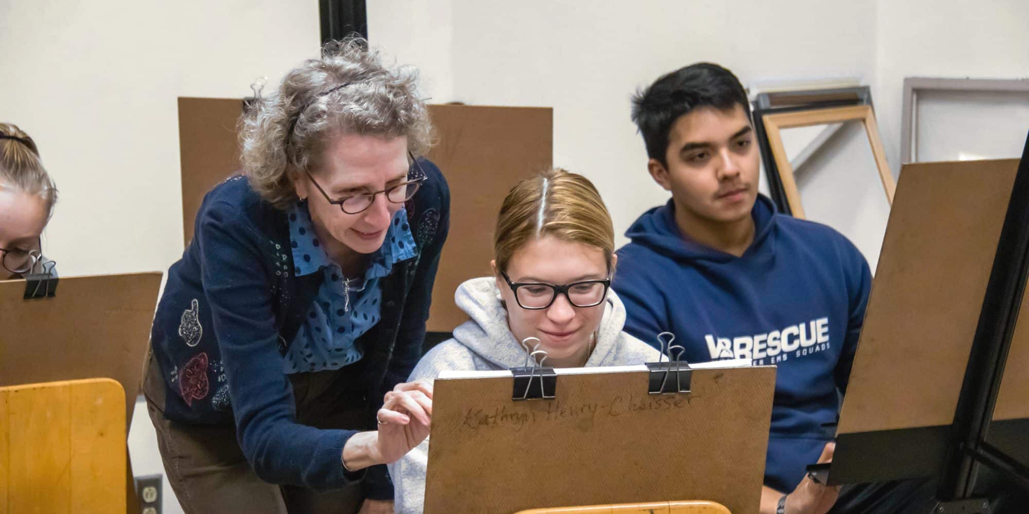 In the art studio, an instructor guides a student working on a drawing, while another student looks on with interest.
