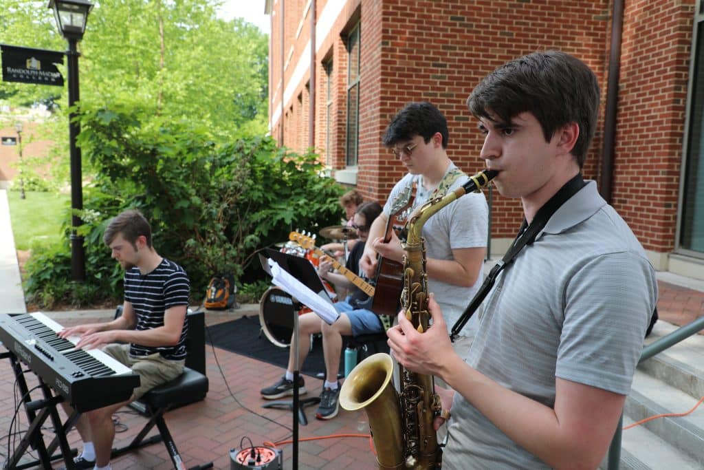 students playing music (sax, piano, guitar, drums) outside a building