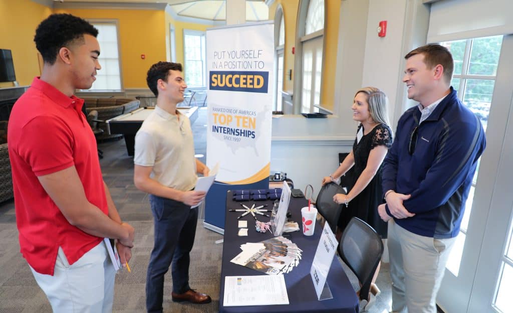 Two students stand speaking to two employers at a table during the Edge Career and Internship Fair