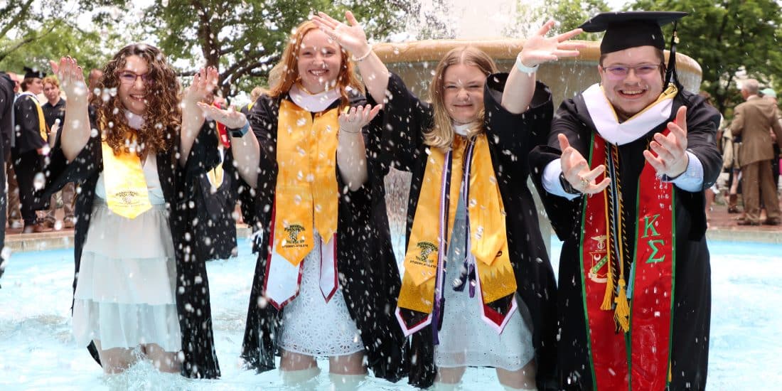 Four students in regalia stand in the fountain splashing water at the camera.