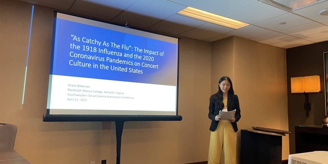 Grace Bakeman reads from her research paper in front of a projector screen.