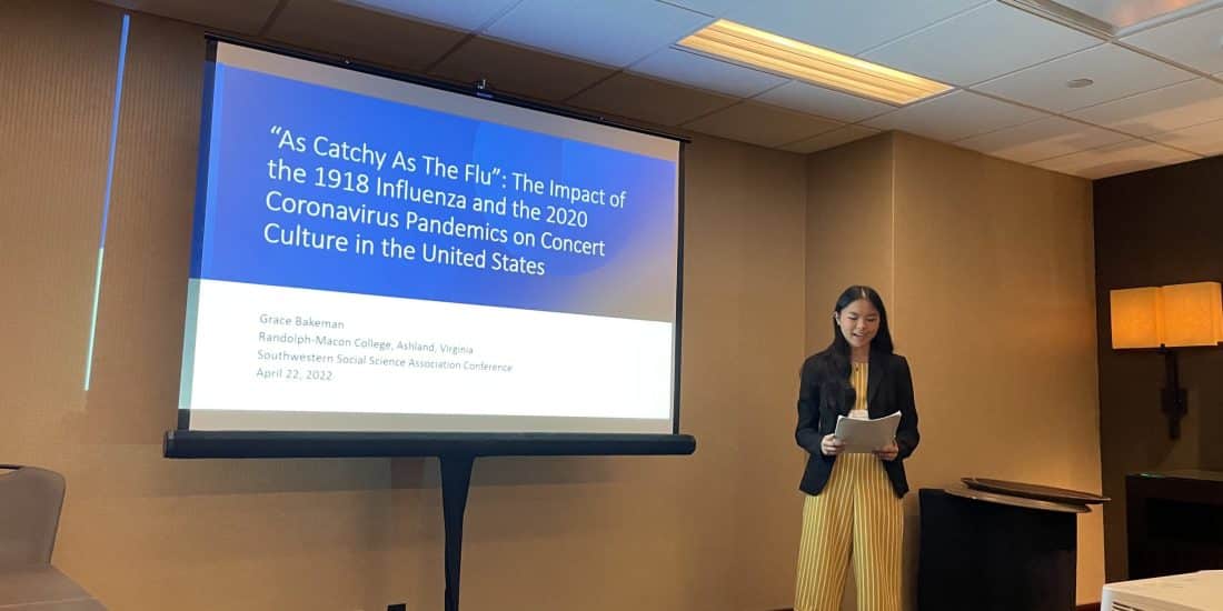 Grace Bakeman reads from her research paper in front of a projector screen.