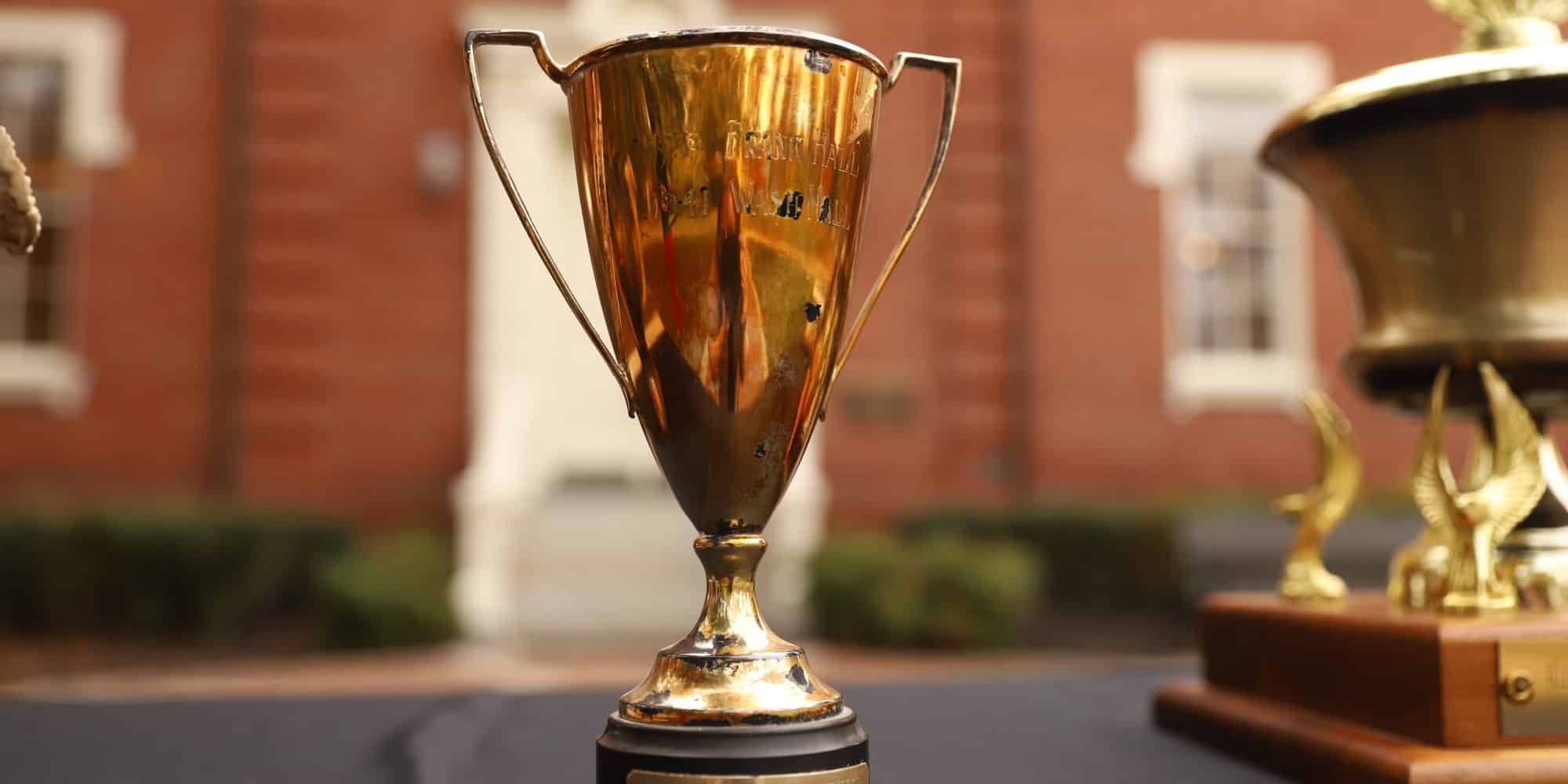 Two gold trophies sit on a table in front of a Wash Frank