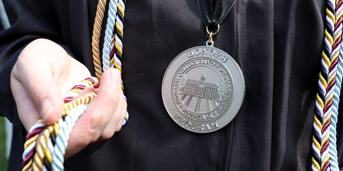 A graduate in a black gown displays their cords and Honors Program medal.