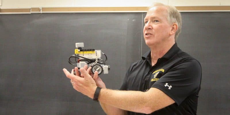 A man holding a small robot in front of a blackboard.