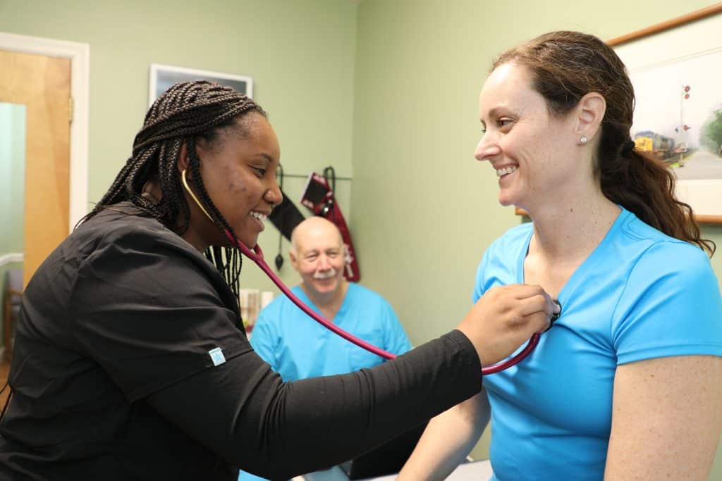 A PA student checks a patient with a stethoscope under the supervision of a doctor
