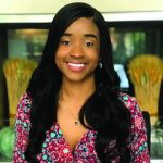 A smiling woman with long wavy hair, wearing a paisley print top, in front of a decorative interior background.