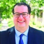 A smiling man in glasses wearing a dark suit and tie.