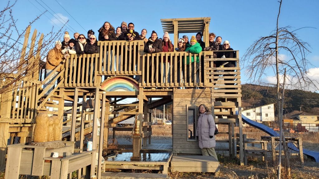 Group of people posing on a wooden playground with a rainbow decoration, one person standing in front.