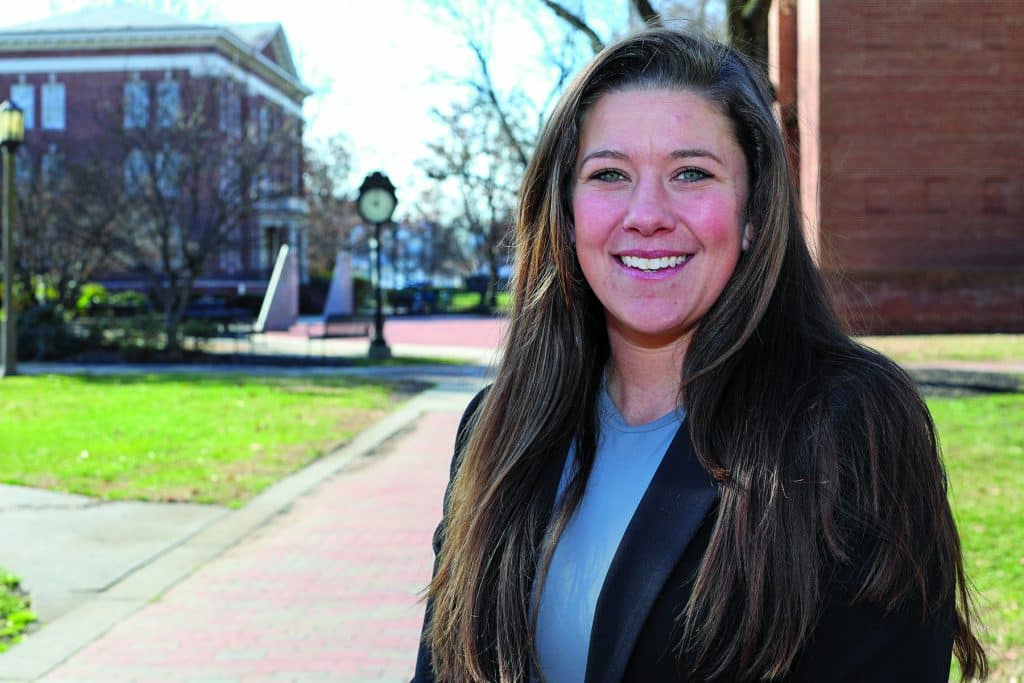 A woman smiling outdoors on a sunny day with buildings and a walkway in the background.