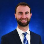 A man in a business suit and tie smiling against a blue background.