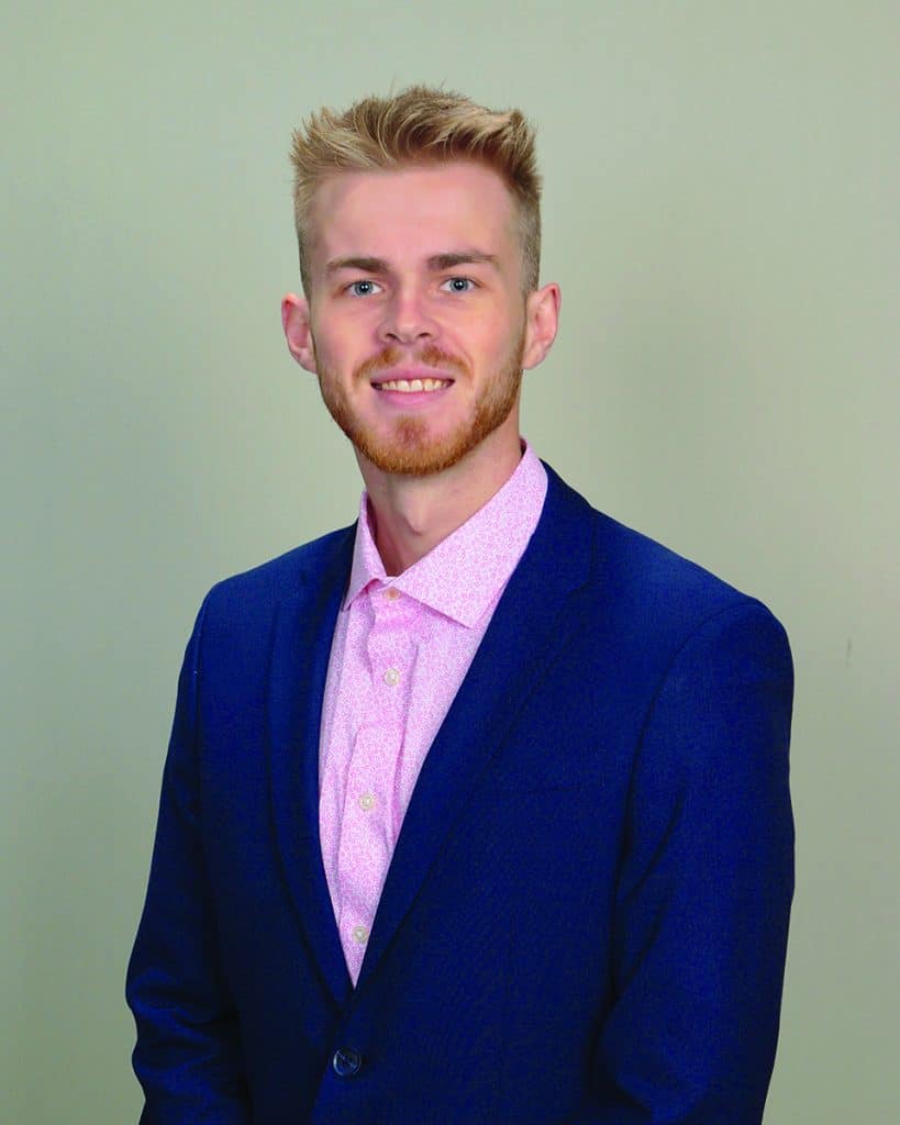 Professional young man in a blue suit with a pink shirt smiling for a portrait.