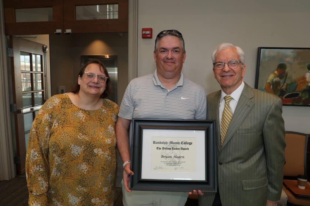 Three individuals posing for a photo, with the middle person holding a framed certificate.