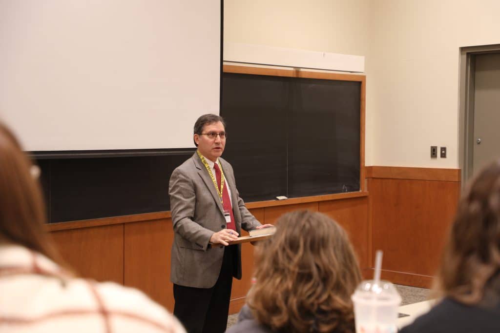 A middle-aged man in a suit lecturing in a classroom while holding a book, with students in the foreground listening.