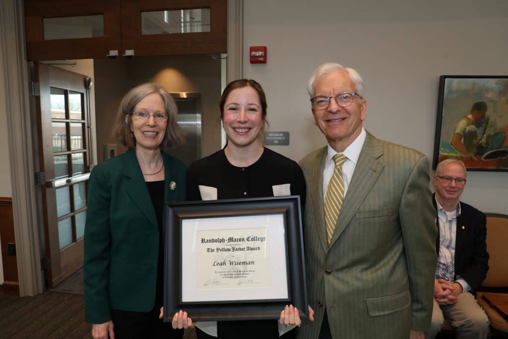 A woman holding a framed certificate stands between two smiling individuals at an award ceremony.
