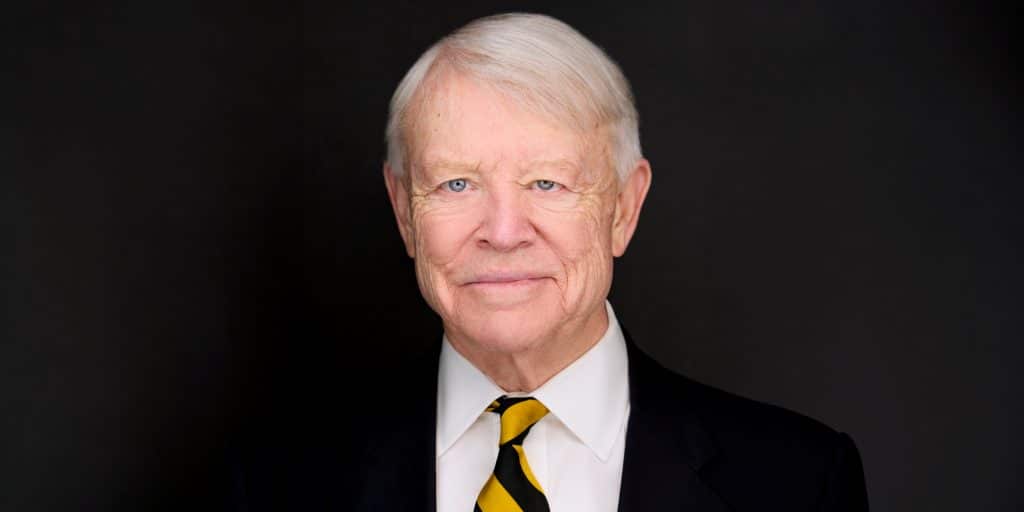 Elderly man with white hair, wearing a black suit and striped tie, smiling gently against a dark background.