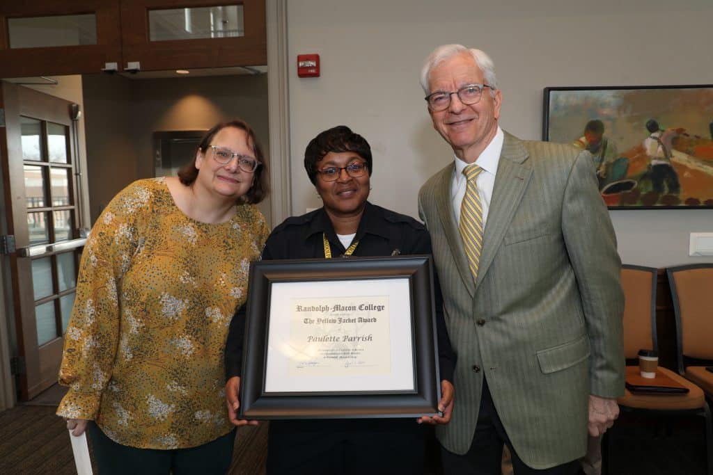 Three individuals smiling at the camera, with the person in the center holding a framed certificate, in an indoor setting.