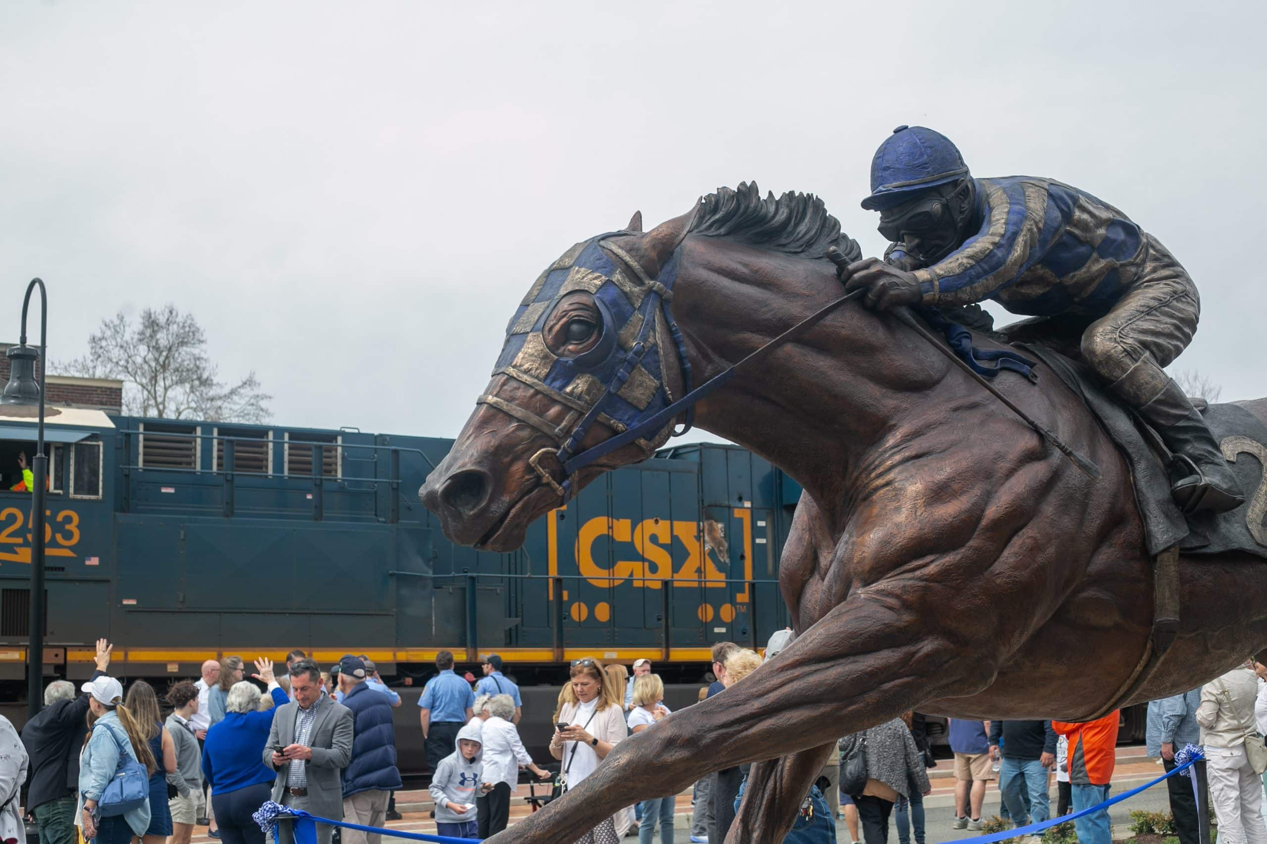 Bronze statue of a jockey on a racehorse with a train passing by in the background and people milling around.