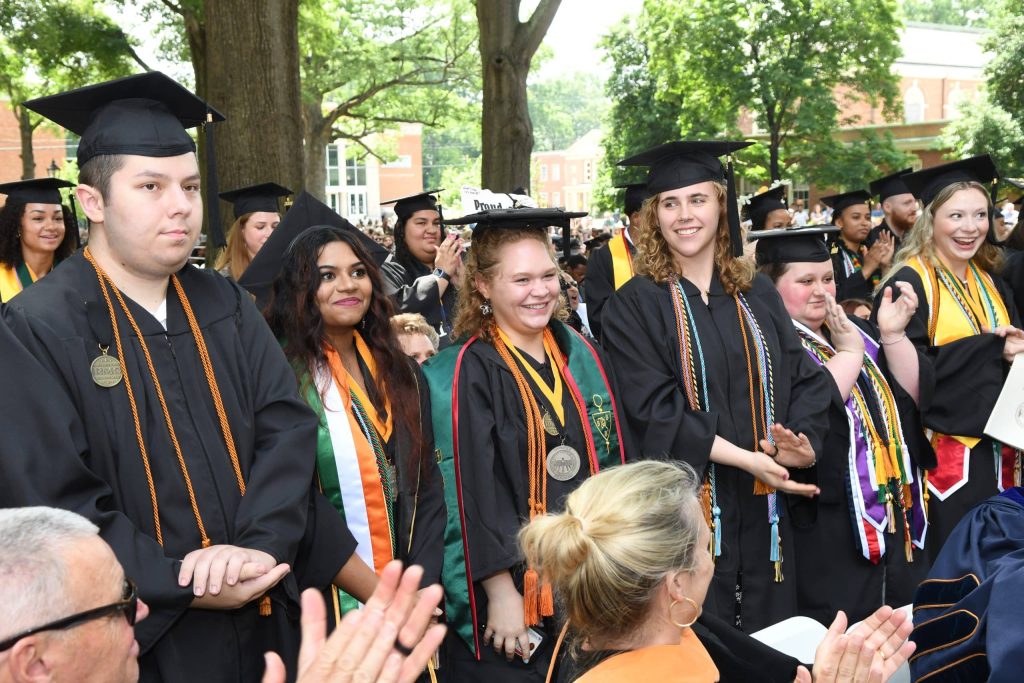 Graduates in caps and gowns stand in a row during a ceremony. Some wear honor cords and medals, and they are surrounded by applauding attendees and greenery.