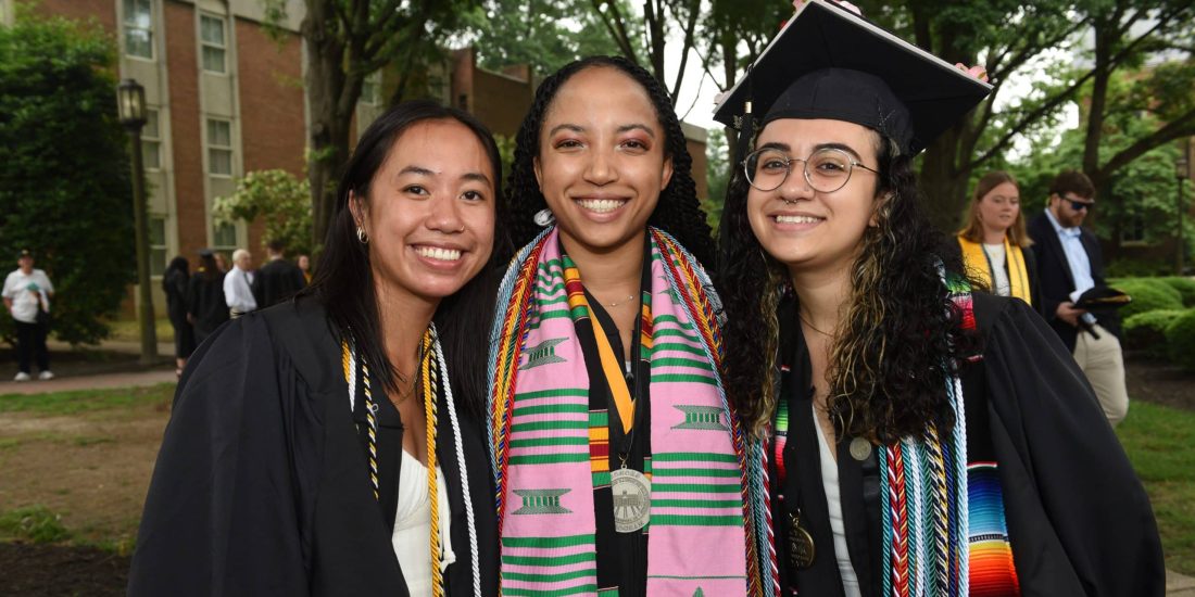 Three graduates in caps and gowns stand side by side, smiling at the camera. They are wearing various honor cords and stoles over their gowns. A building and trees are visible in the background.