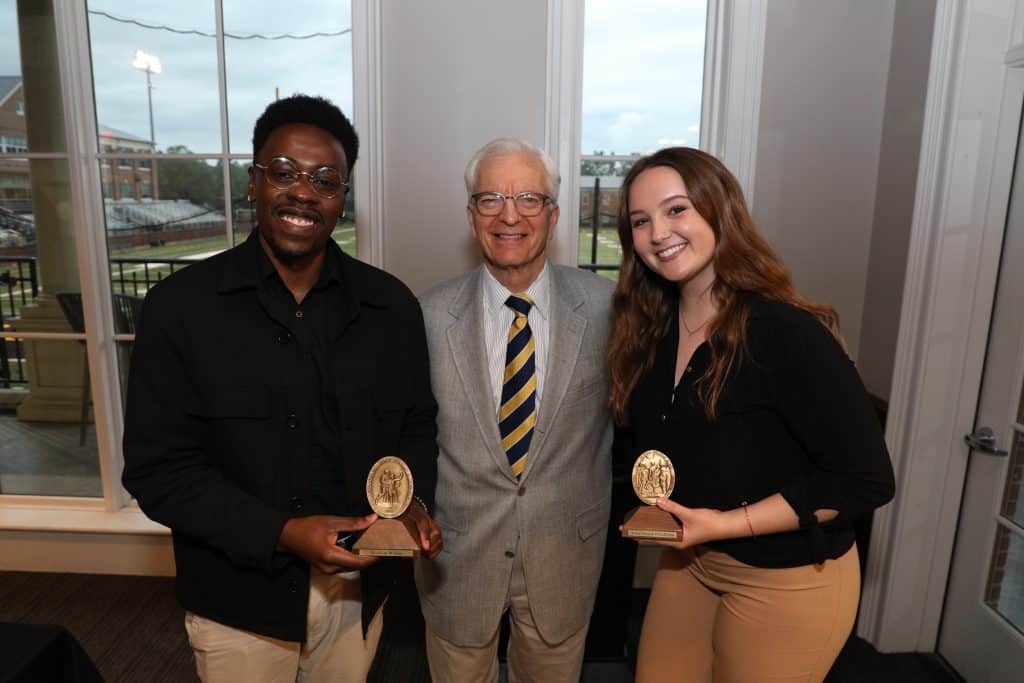Two students and an older man holding awards, smiling at the camera in a room with large windows showing a sports field outside.