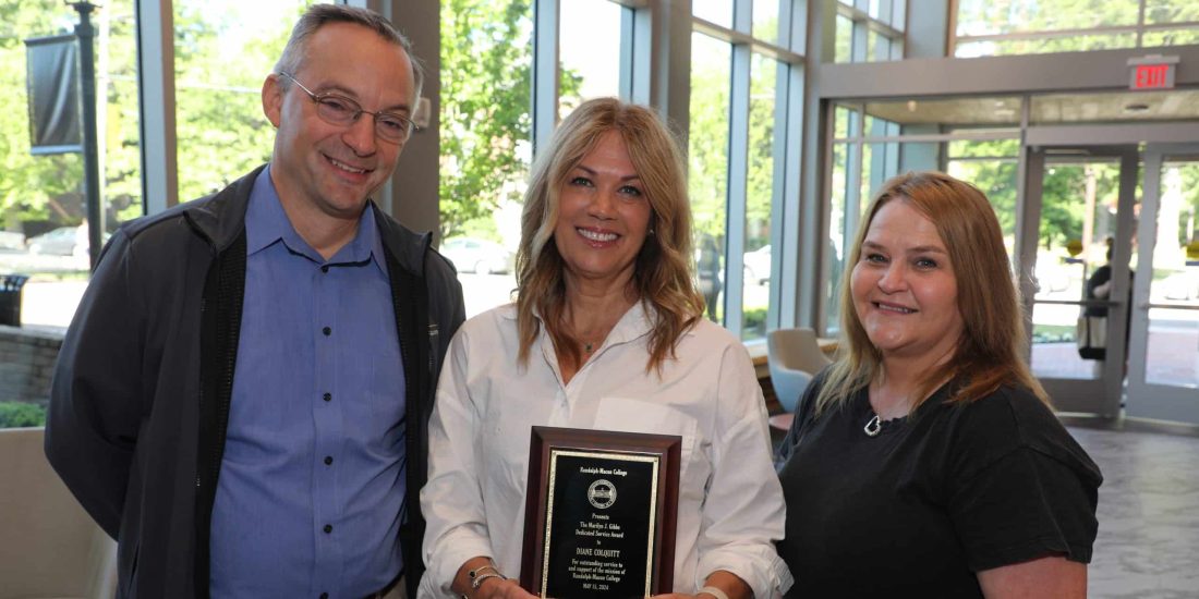 Three people stand indoors near large windows. The person in the middle holds a plaque, smiling. The other two are also smiling, standing on either side.