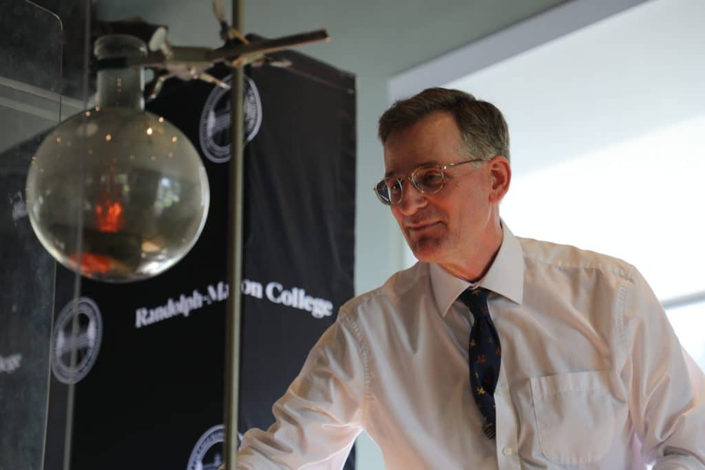 A man in a white shirt and tie observing a scientific demonstration involving a suspended glass sphere.
