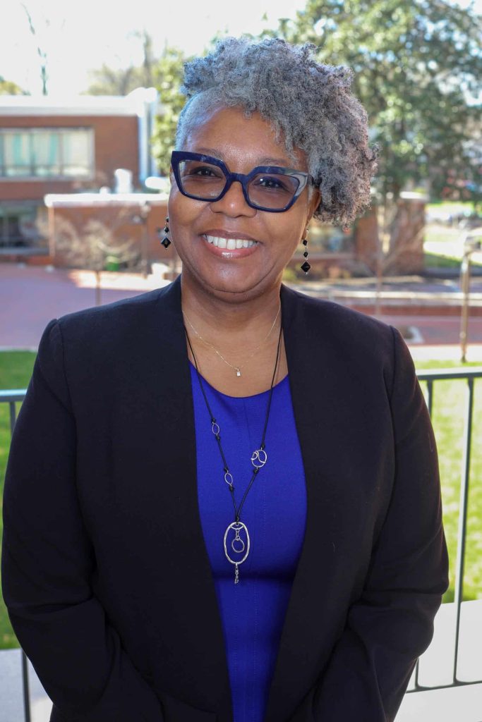 A woman wearing glasses and a blue dress with a black blazer smiles while standing outdoors in front of a building.