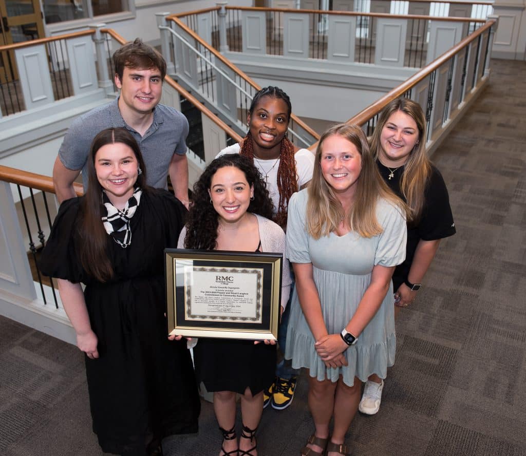 Six students standing on stairs, displaying an award plaque, smiling at the camera in an indoor setting.